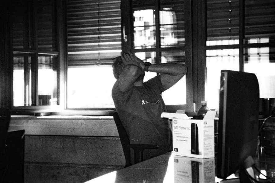 Black-and-white photo of someone at a desk, hands on their head, facing a monitor