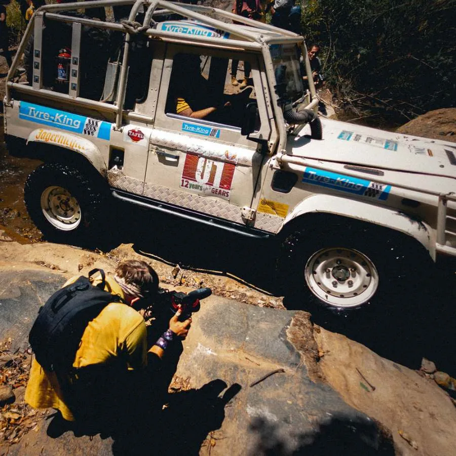 Photographer crouched on a rock filming a rally Land Rover as it climbs over